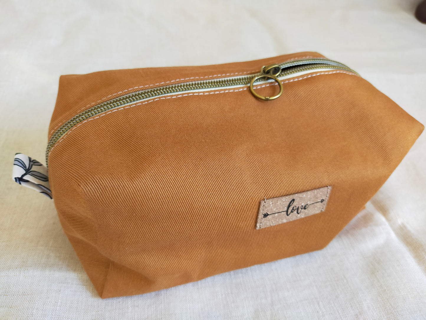vue de dessus, trousse de toilette couleur ocre, avec fermeture éclaire bronze et étiquette en cuir recyclé apposé sur l'avant, notée Love