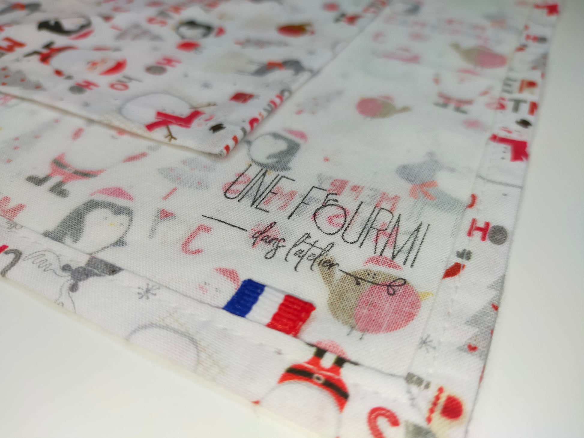 zoom sur les coutures propres du furoshiki avec le tampon de la marque une fourmi dans l'atelier et le ruban tricolore inséré dans la couture. tissu en coton thème noel avec père noel et pingouins